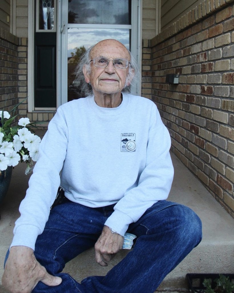 Guillermo sits in jeans and a crewneck sweatshirt in front of a brick entryway with potted flowers