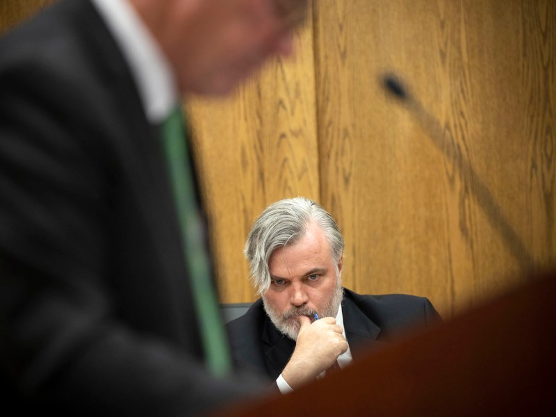 Jay Jerde appears beyond a blurry speaker at a podium. Jerde is pensive, staring into the middle distance with a thumb on his lips and a pen in his hand
