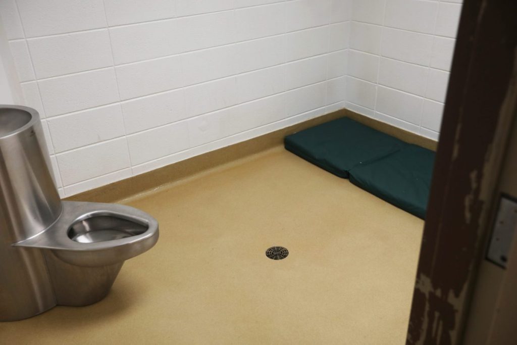 A small room with just a metal toilet and sleeping pad, with a drain in the middle