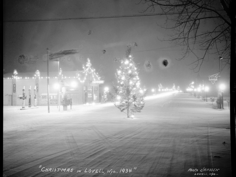 A black and white photo of downtown Lovell. There's a thin layer of snow showing where cars had driven earlier that day, Christmas lights on a few shops, bright round street lights, and a lit-up tree right in the middle of the road.