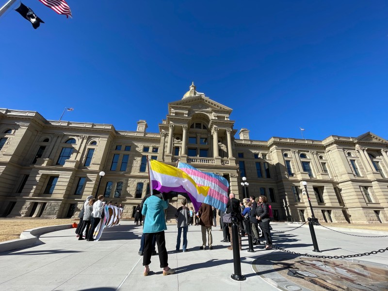 People hold the trans pride flag and the gender nonbinary pride flag in front of the Wyoming Capitol building on a sunny, windy day