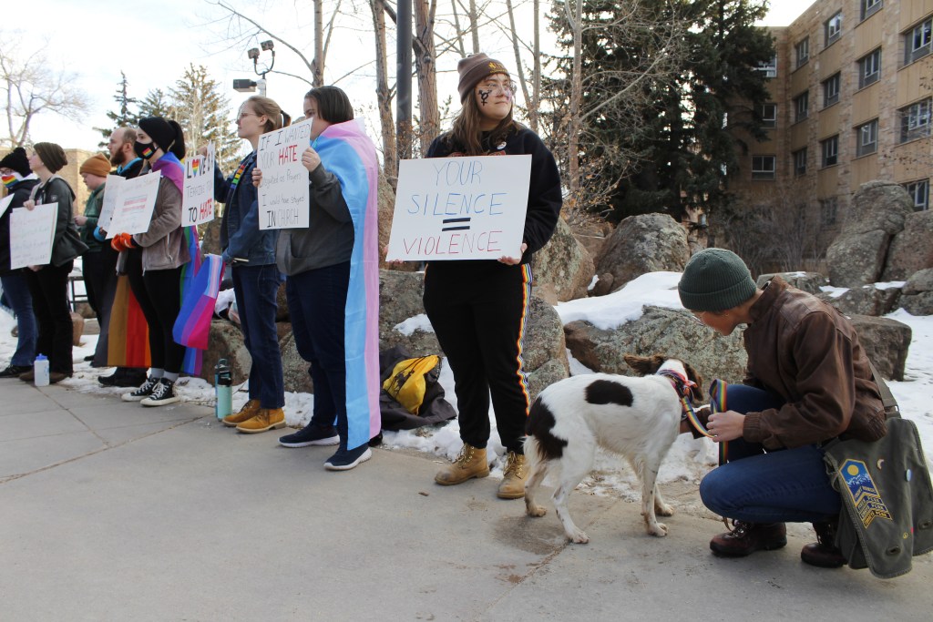 UW students stand in a line holding pro-LGBTQ signs that say "your silence = violence" and "If I wanted your hate, I would've stayed in church." There is also a dog with a rainbow leash