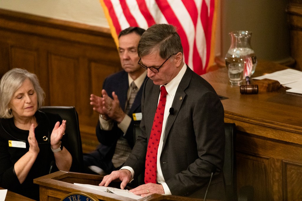 Mark Gordon stands behind a podium looking down while people clap in the background