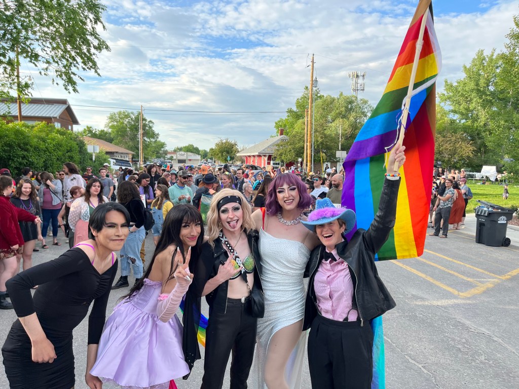 A group of drag queens and a drag king smile and hold a rainbow flag