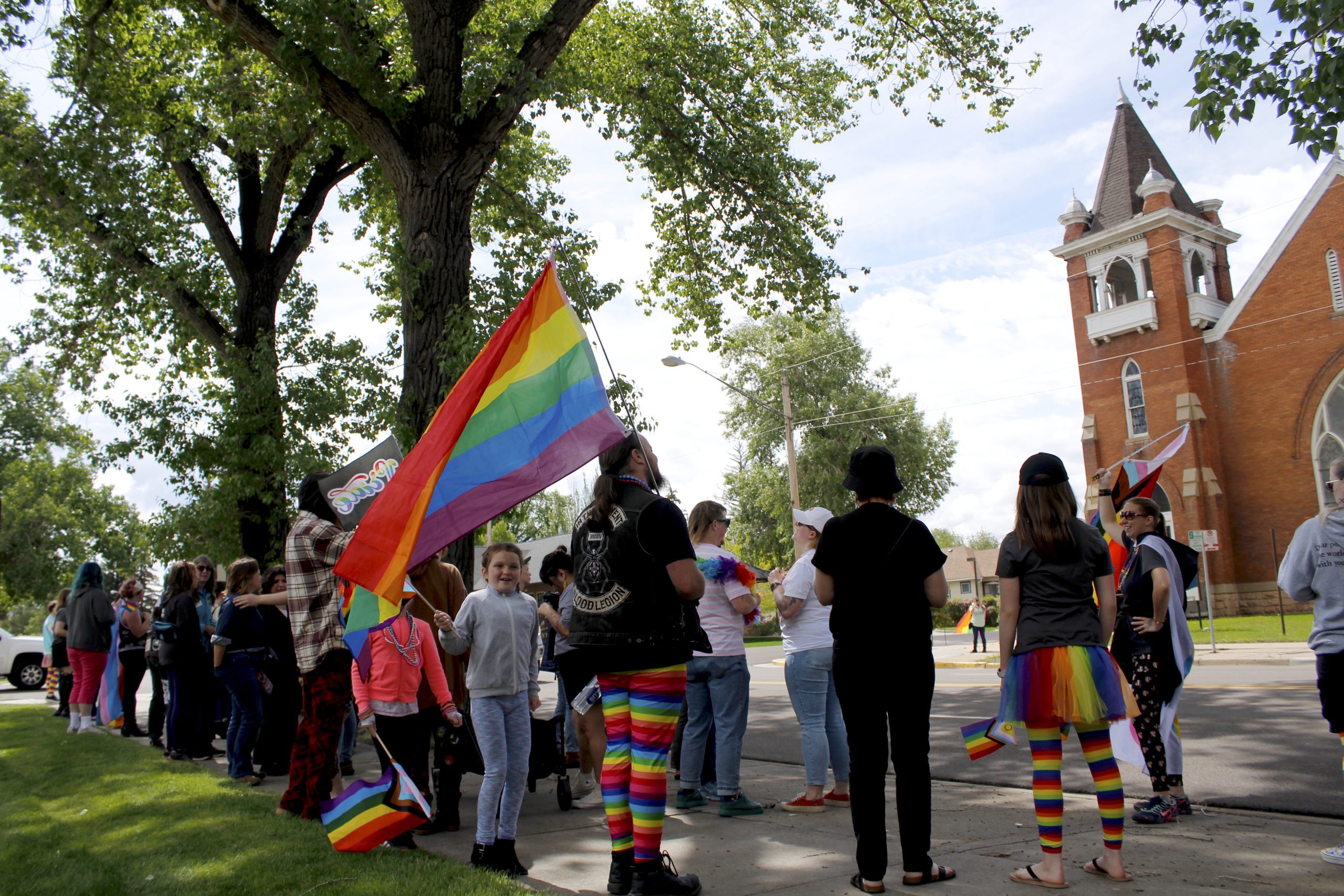 Families with pride flags and colorful clothing stand beside Grand Ave in front of the Laramie Counthouse