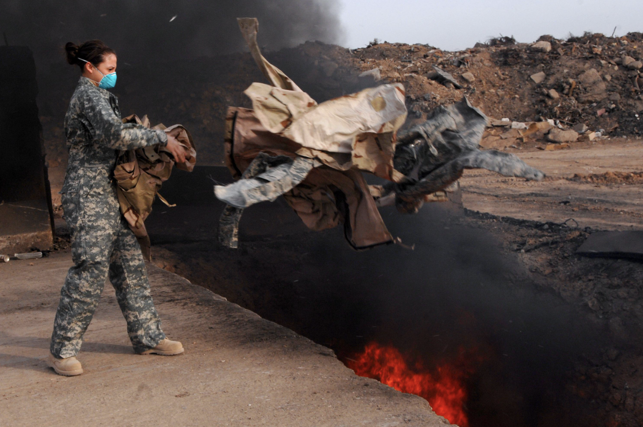 A woman with a surgical mask throws clothes into a deep burning pit with black smoke wafting up behind her