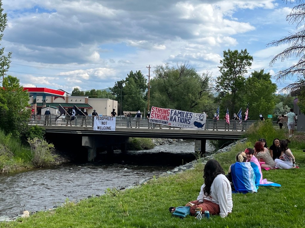 Patriot front members with neck gaters, American flags and signs stand on the bridge in Lander as Pride celebrants look on