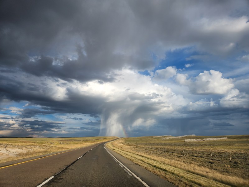 An empty road leads to a microburst with snow fencing and yellow grasses along the roadside. There is a rainbow ahead as well.