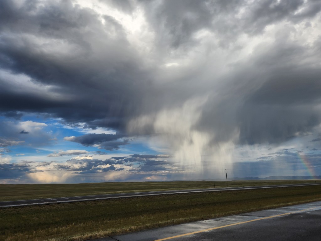 Thin lines streak down faintly from a cloud overlooking the prairie. Fresh rain can be seen glistening off of the roadway