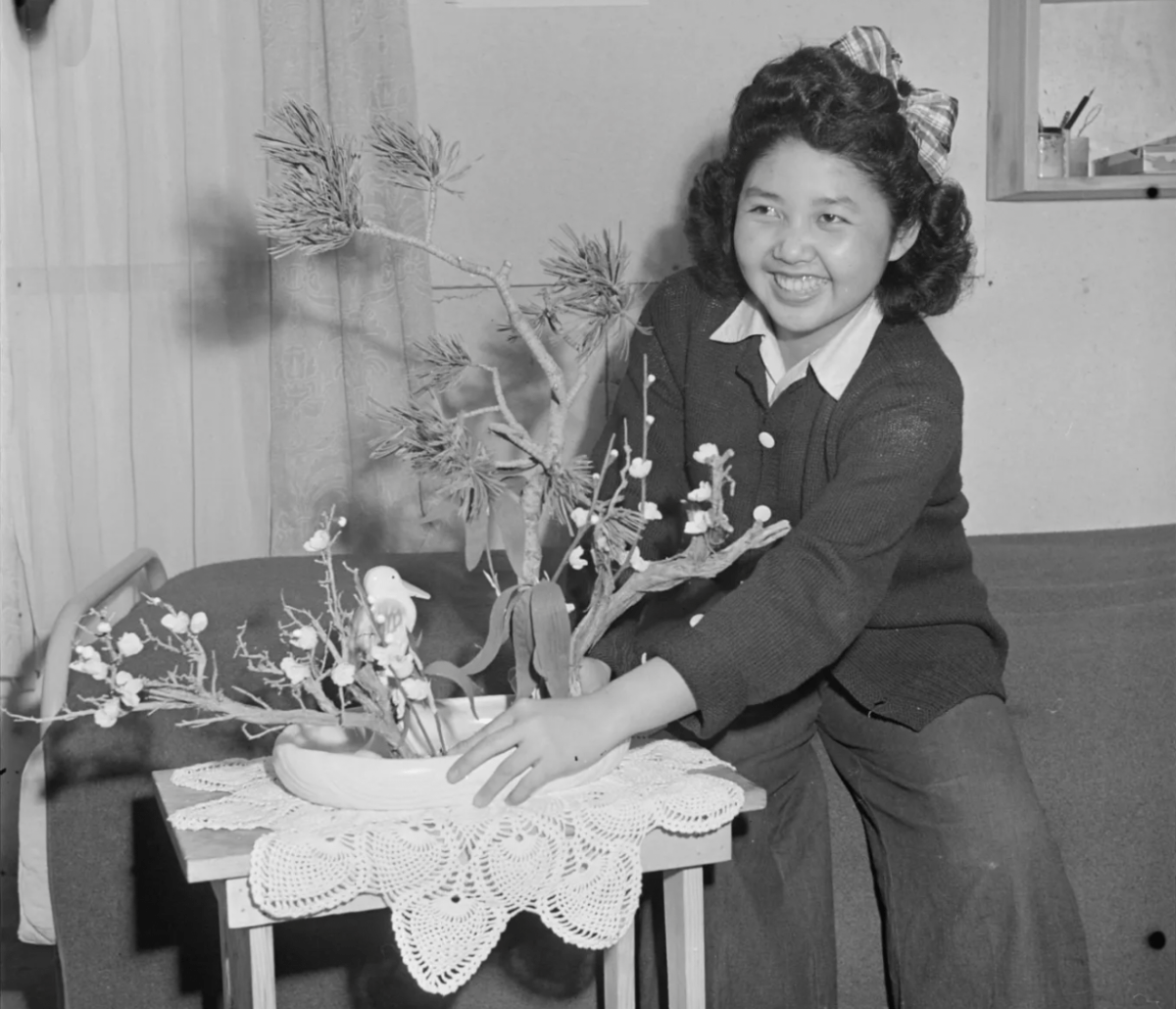 A young girl with curly hair, sweater and bow proudly displays a New Year's arrangement with a smile on her face. The arrangement is simple, with three visible branches and a small bird decoration