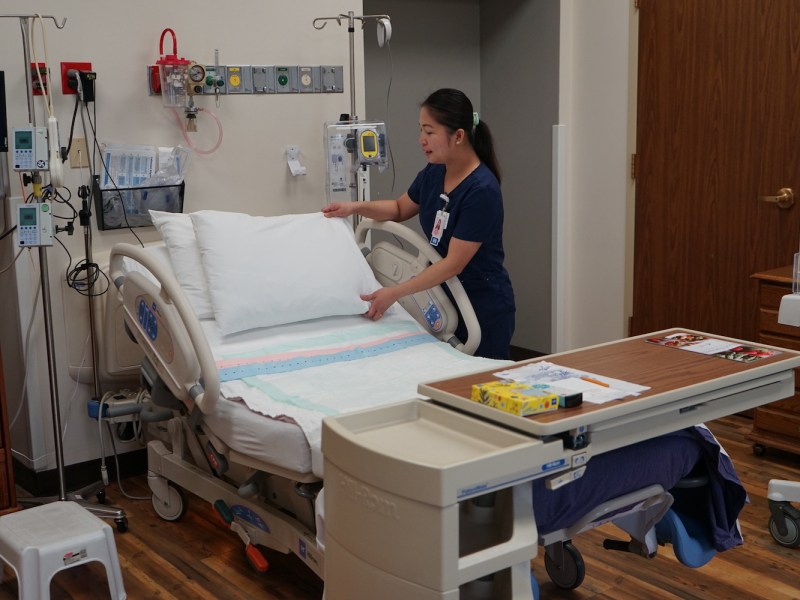 A woman in scrubs places a pillow on a hospital bed