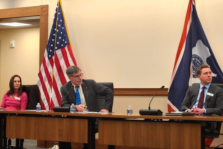 Three people sit behind a wooden table, looking off to the right