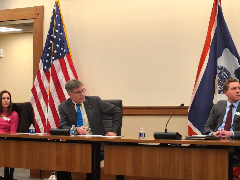 Three people sit behind a wooden table, looking off to the right