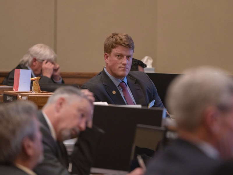 A man in a suit looks over others' head on the House floor, straight into the camera