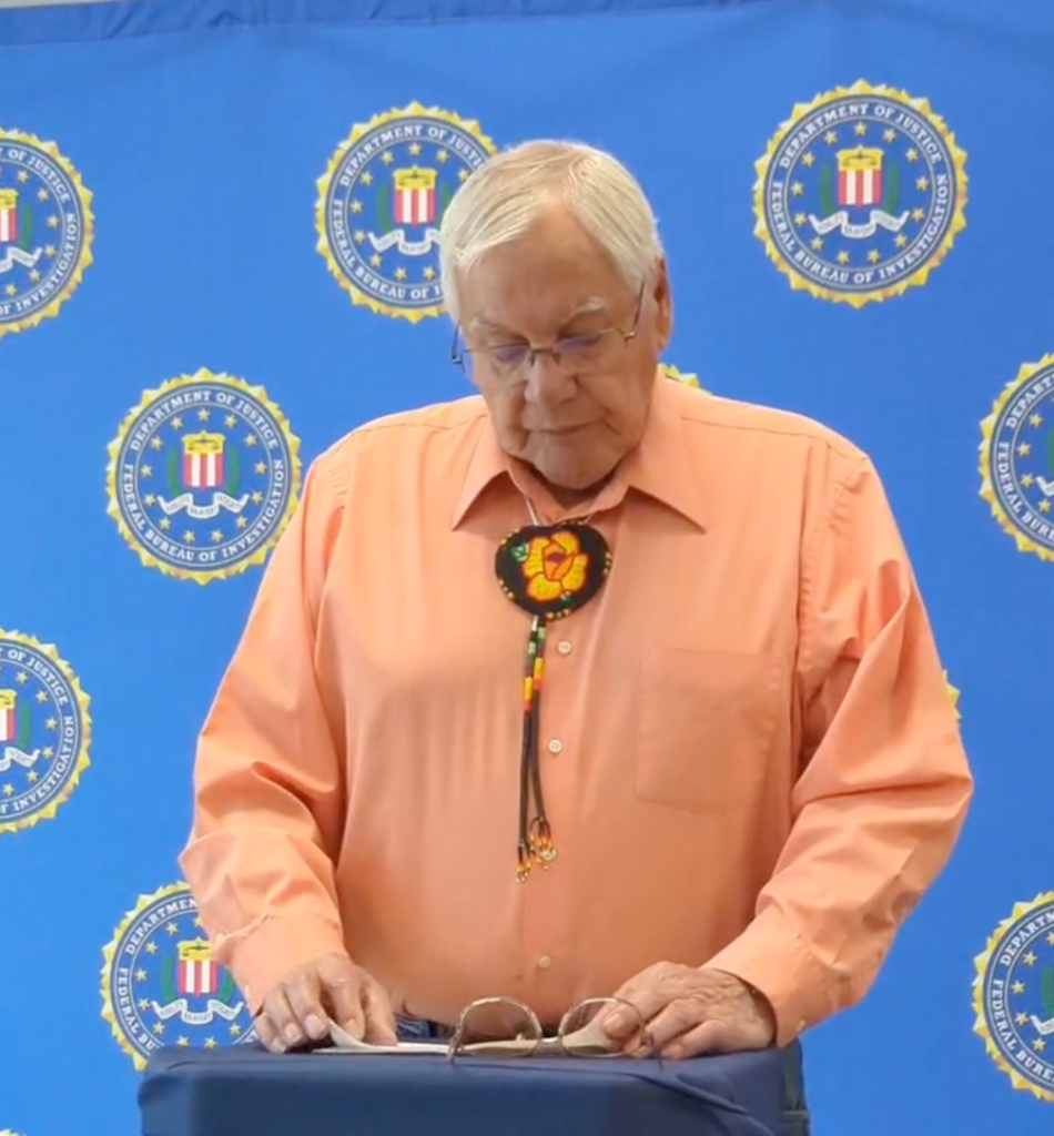 A man stands at a podium in front of a blue wall with FBI logos