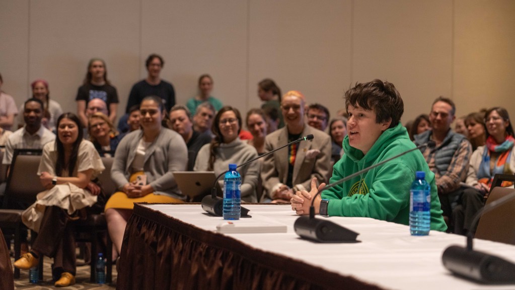 Lauren McLane sits at a table speaking into a microphone while a crowd looks on