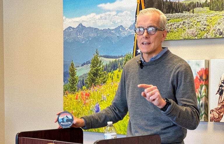 Secretary Denis McDonough stands at a podium, speaking in front of pictures of Wyoming wilderness