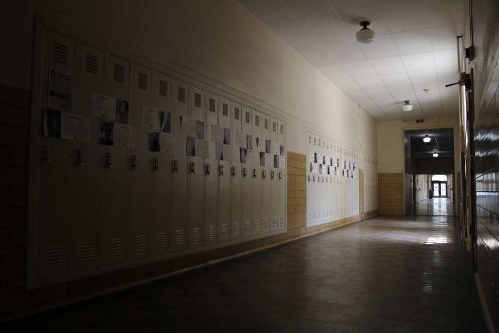 A hallway with patchy sunlight shining through windows onto old lockers that have unissued diplomas taped to them