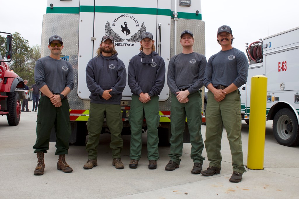 Five men in grey shirts and green pants stand in a line