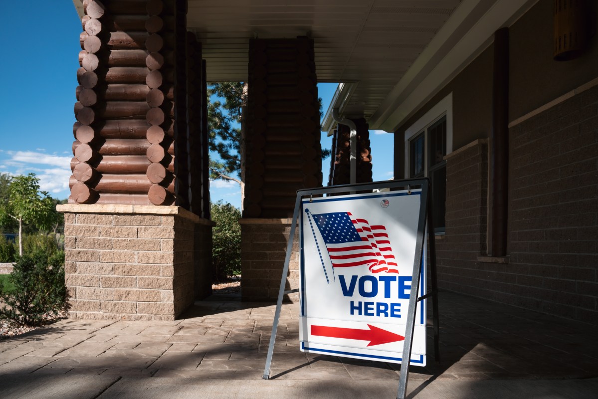 A "vote here" sign with an arrow and American flag in front of a brick building