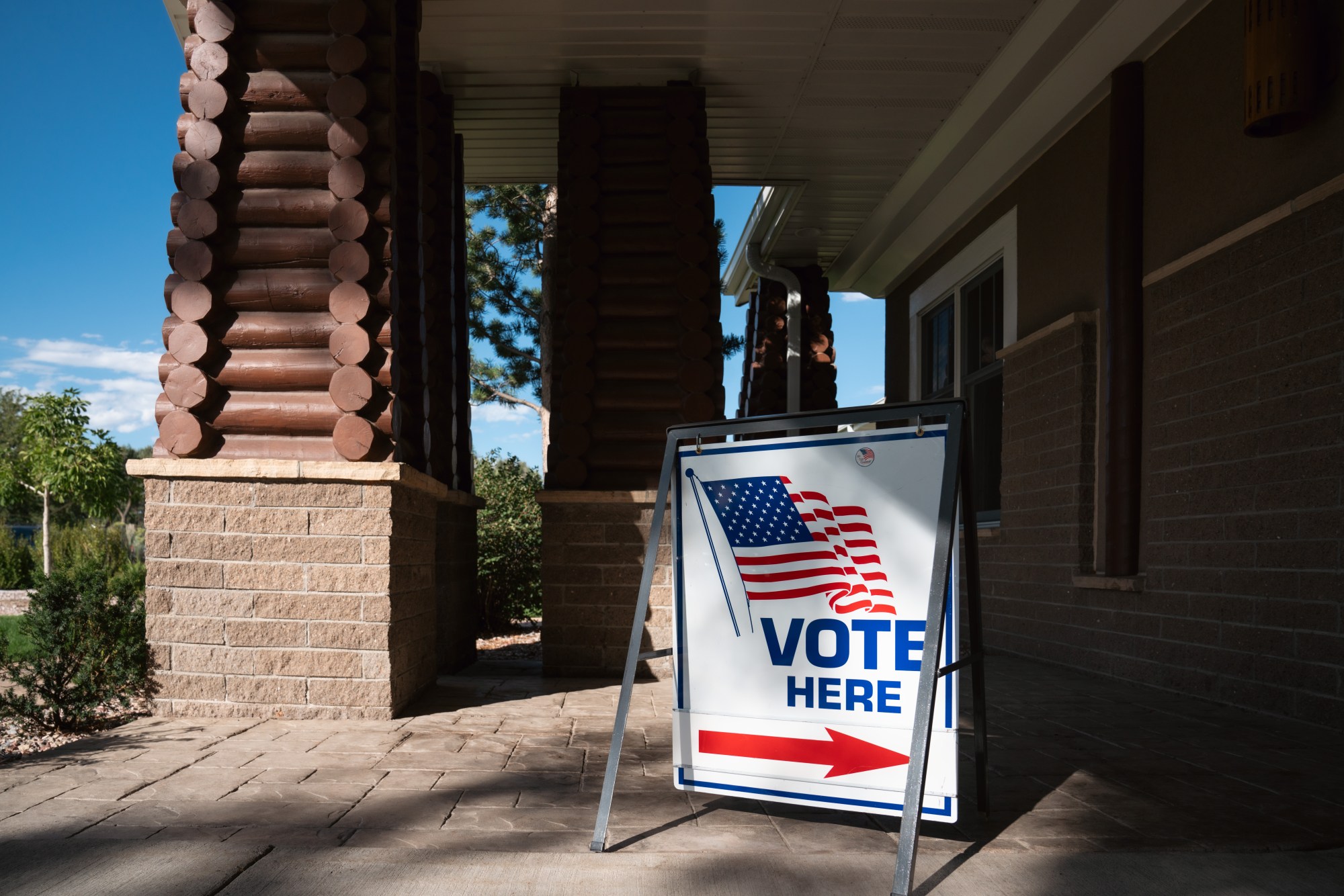 A "vote here" sign with an arrow and American flag in front of a brick building
