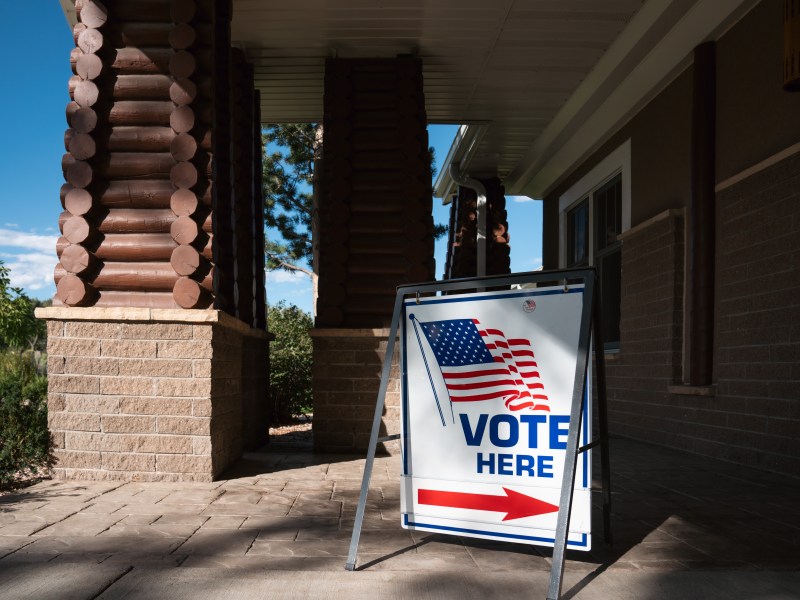 A "vote here" sign with an arrow and American flag in front of a brick building