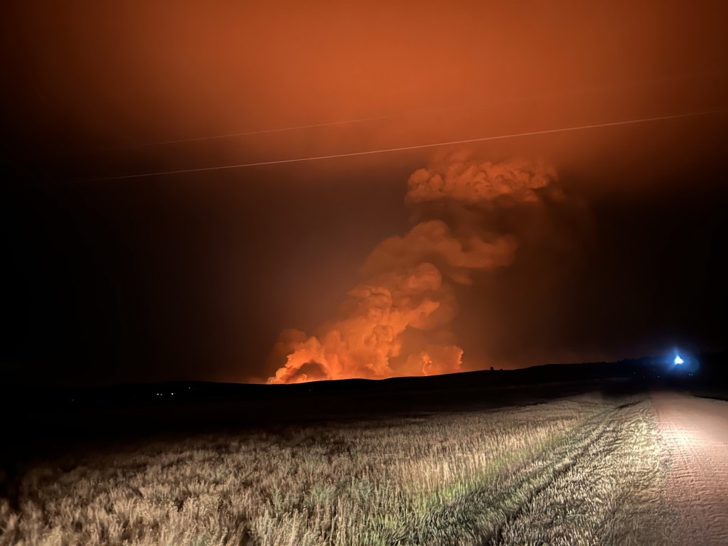 The orange glow of a fire burning at night. It's clearly taken from a dirt road near a vehicle with its lights on