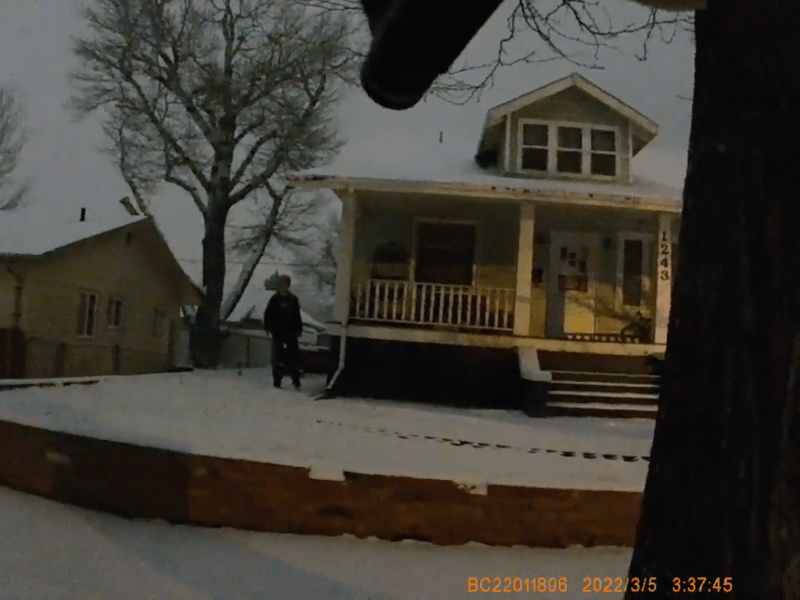 A man stands in a snowy yard while a firearm barrel can be seen pointing towards him from the vantage of the bodycam wearer