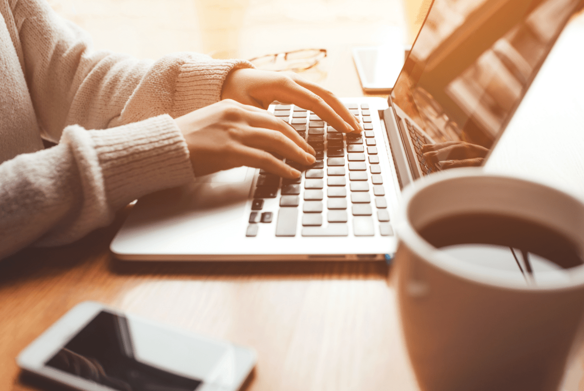 Hands typing on a laptop keyboard, with sunlight streaming in. A cup of coffee and a smartphone are on the wooden table, conveying a cozy, focused mood.