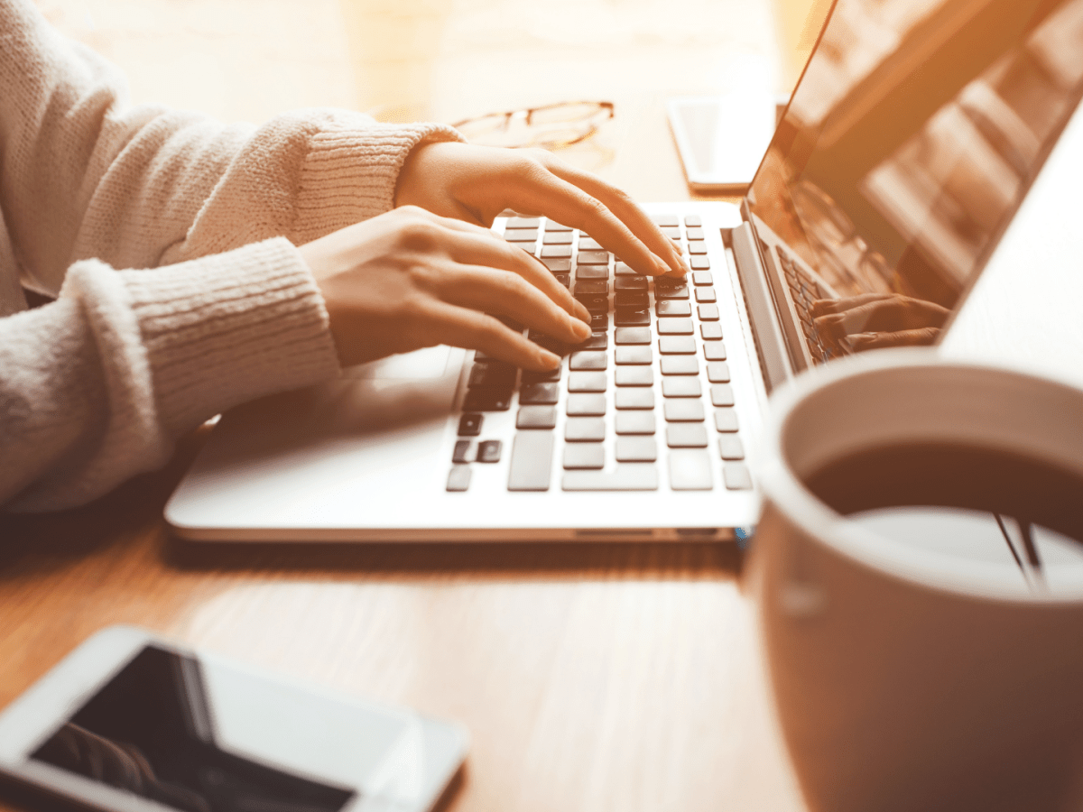 Hands typing on a laptop keyboard, with sunlight streaming in. A cup of coffee and a smartphone are on the wooden table, conveying a cozy, focused mood.